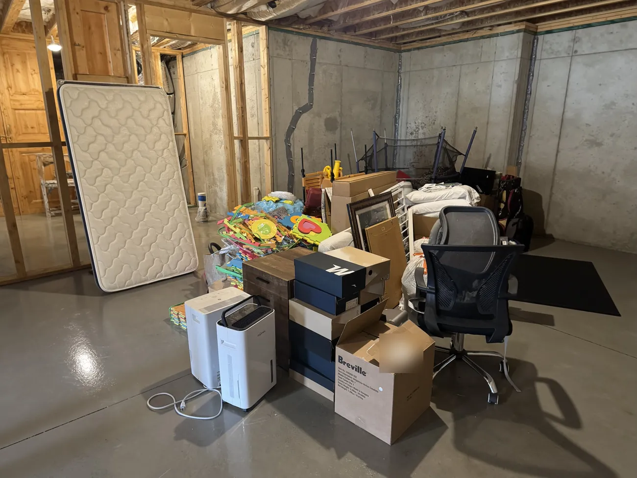 A basement with a mattress, boxes, an office chair, two dehumidifiers and children's play mats stacked up before a cleanout