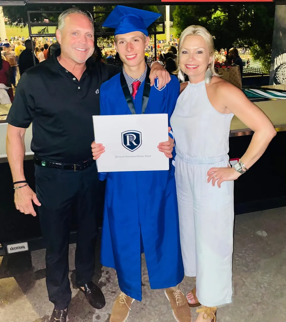 A graduate in a blue cap and gown holding a diploma, standing between two smiling adults at an outdoor graduation ceremony
