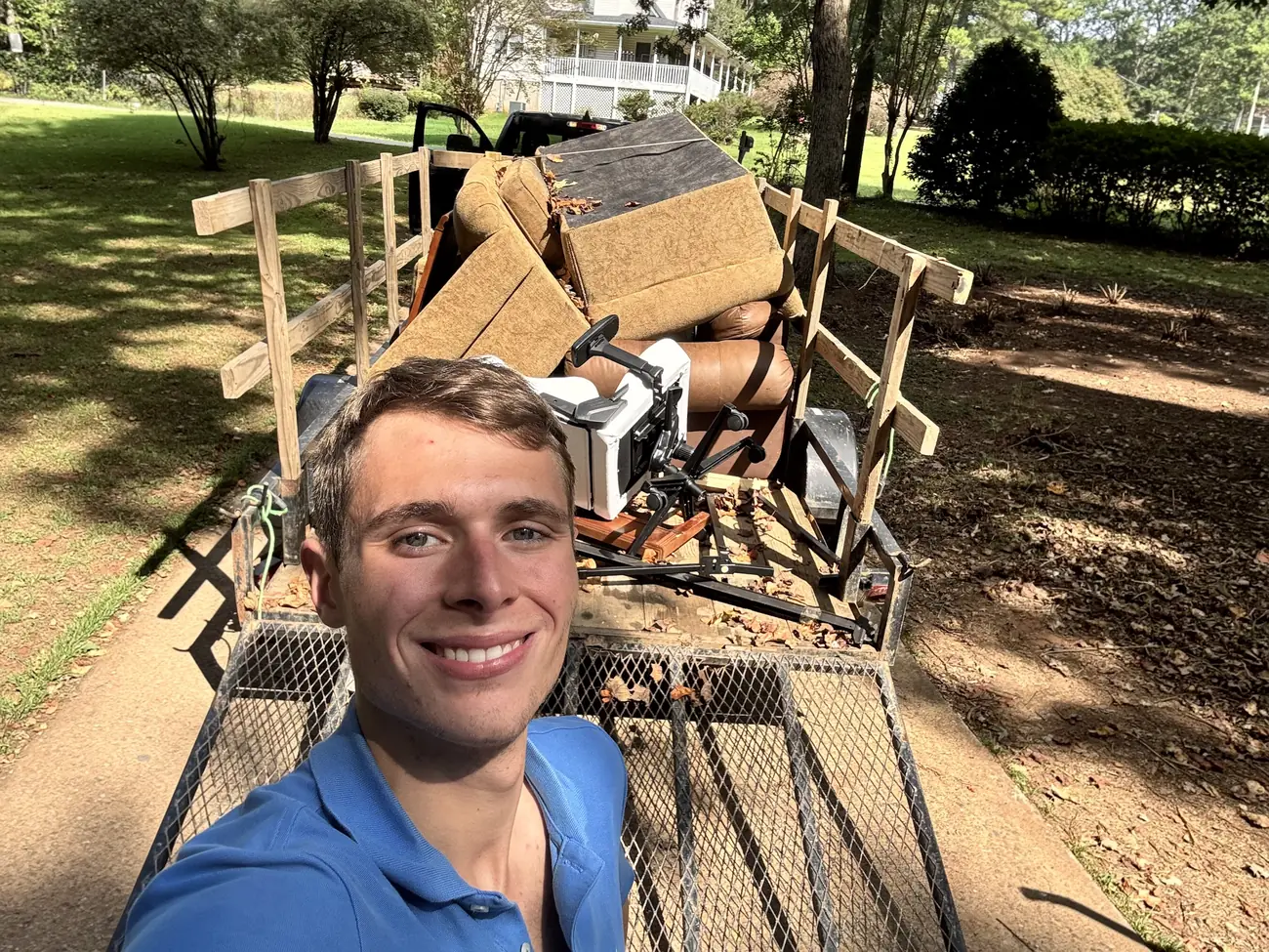 Brayden, owner of Eagle Junk Removal, smiling in front of his trailer loaded with old couches and an office chair